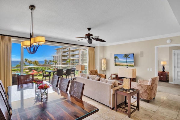 A modern hotel room with a dining area, a sofa set, a ceiling fan, and a large window with a view of the ocean.