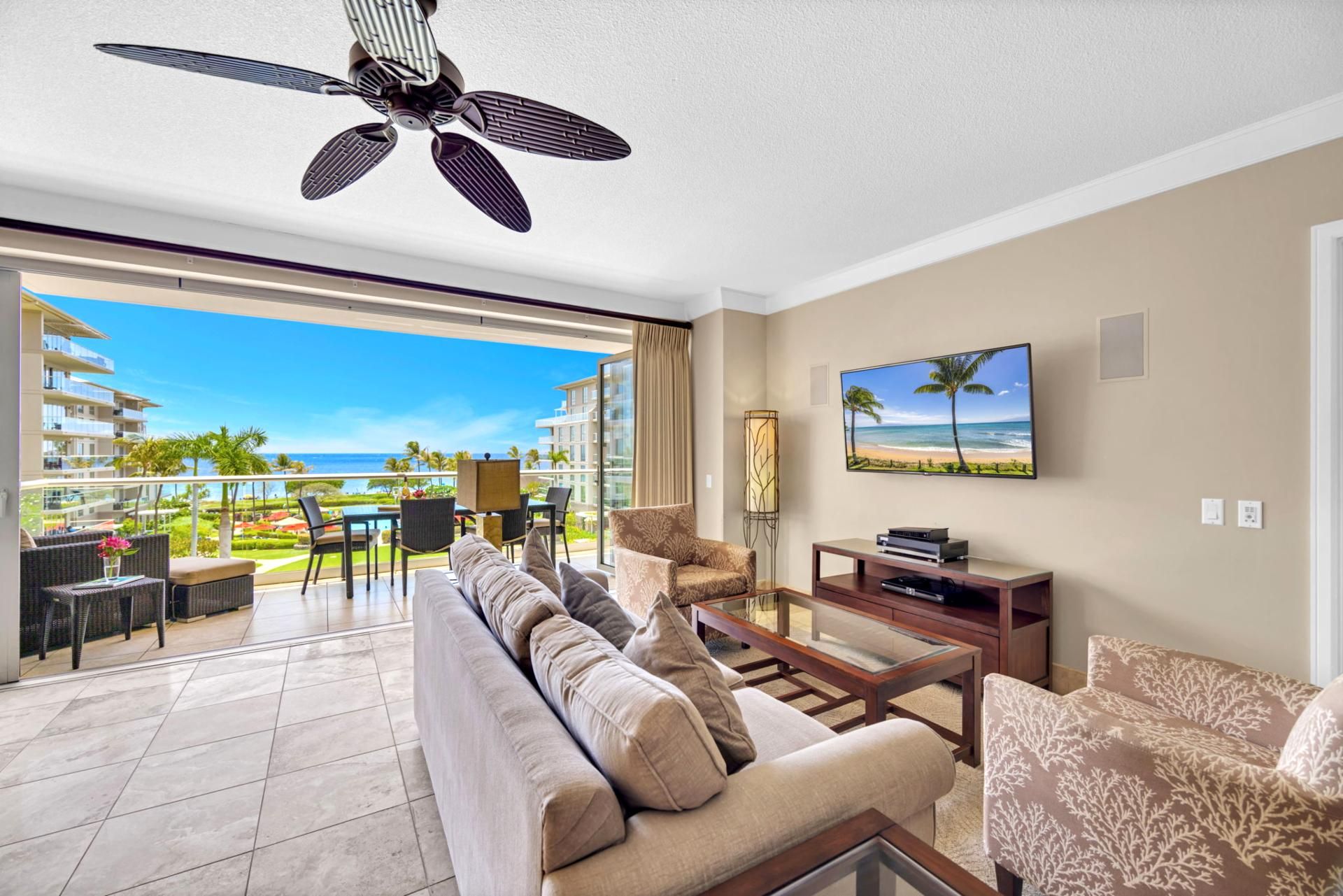 A spacious living room with a large sofa, a coffee table, and a TV mounted on the wall. There is a balcony with a view of the ocean and palm trees.