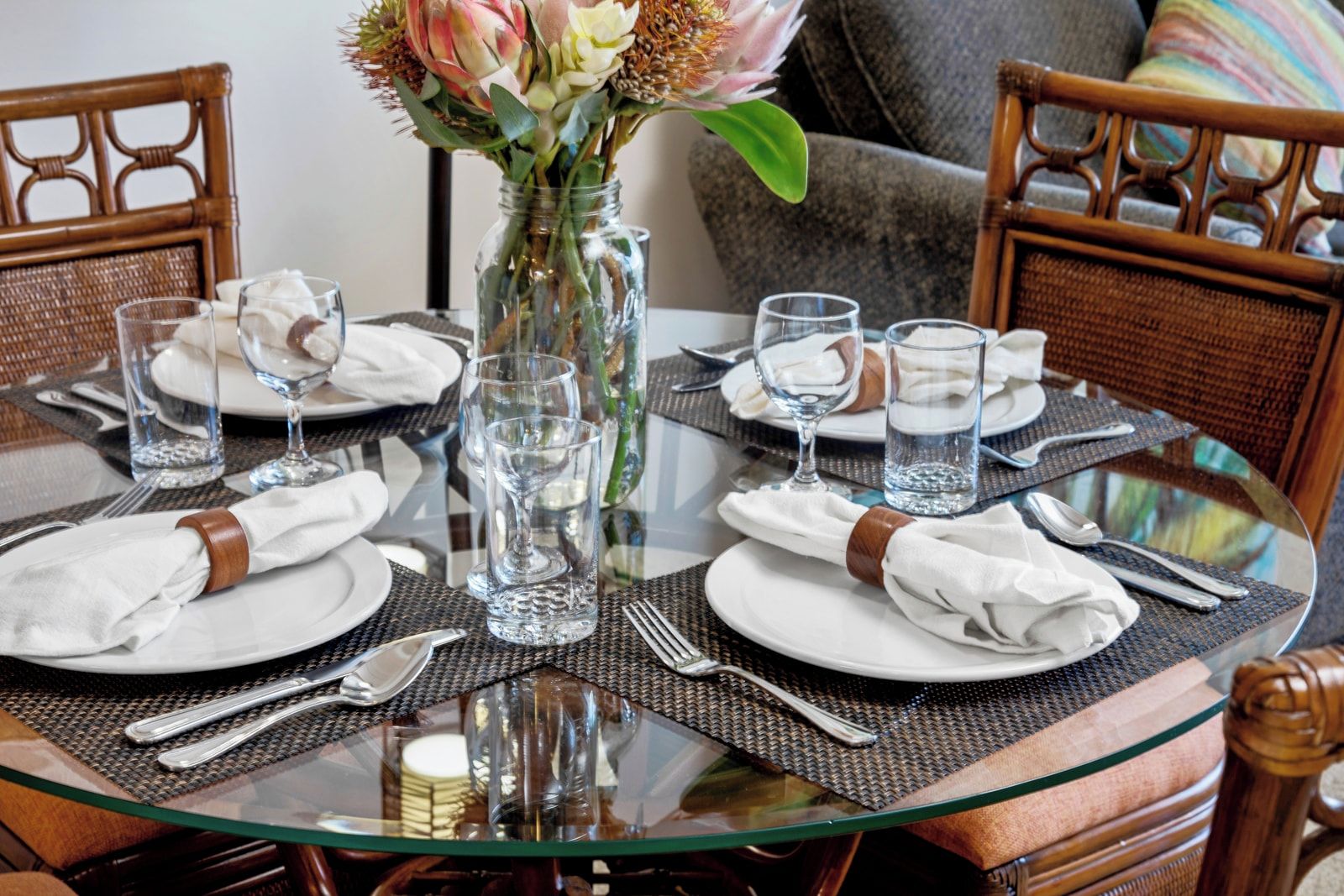 A round glass table set for four with plates, cutlery, and glasses, a vase with flowers, and a wicker chair.