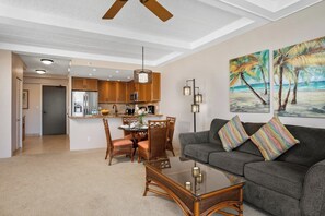 A modern living room with a gray sofa, a dining table with chairs, a kitchen area with wooden cabinets, and a ceiling fan.
