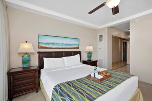 Une chambre d’hôtel avec un lit, deux tables de chevet avec des lampes, un tableau représentant une plage, un ventilateur de plafond et une porte donnant sur une autre pièce.