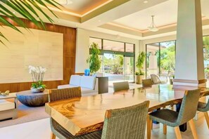 A modern hotel lobby with a large dining table, comfortable seating, and large windows offering a view of the outdoors.