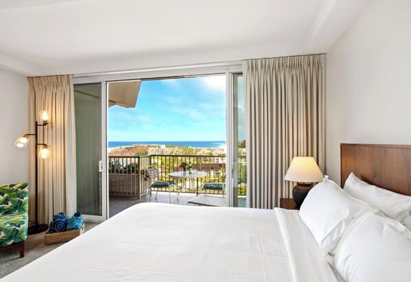 A hotel room with a large bed, a balcony with a view of the beach, a table with chairs, and a lamp.