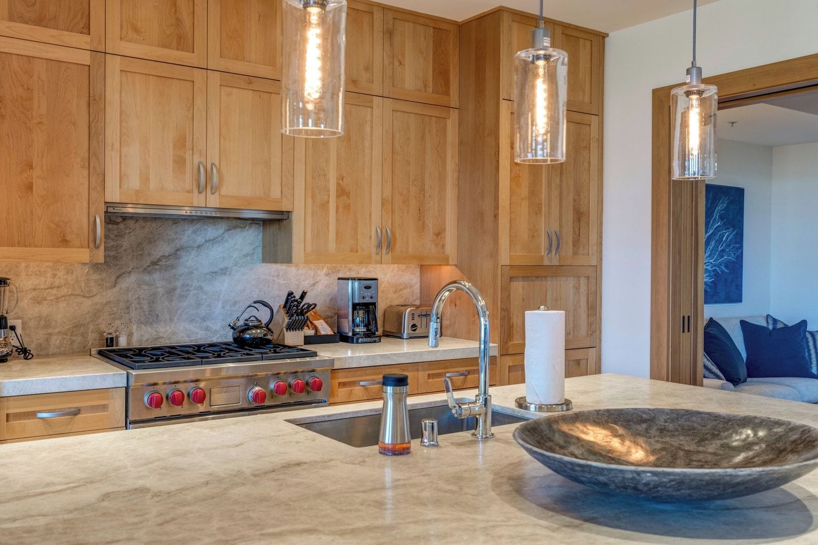 A modern kitchen with wooden cabinets, a gas stove, a coffee maker, and pendant lights.