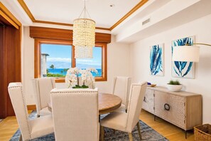 Un coin repas avec une table ronde, quatre chaises, un lustre, une vue sur l’océan et des œuvres d’art accrochées au mur.