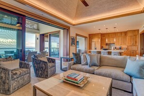 A spacious living area with a sofa, armchairs, and a coffee table. There is a kitchen in the background with wooden cabinets and a dining area.