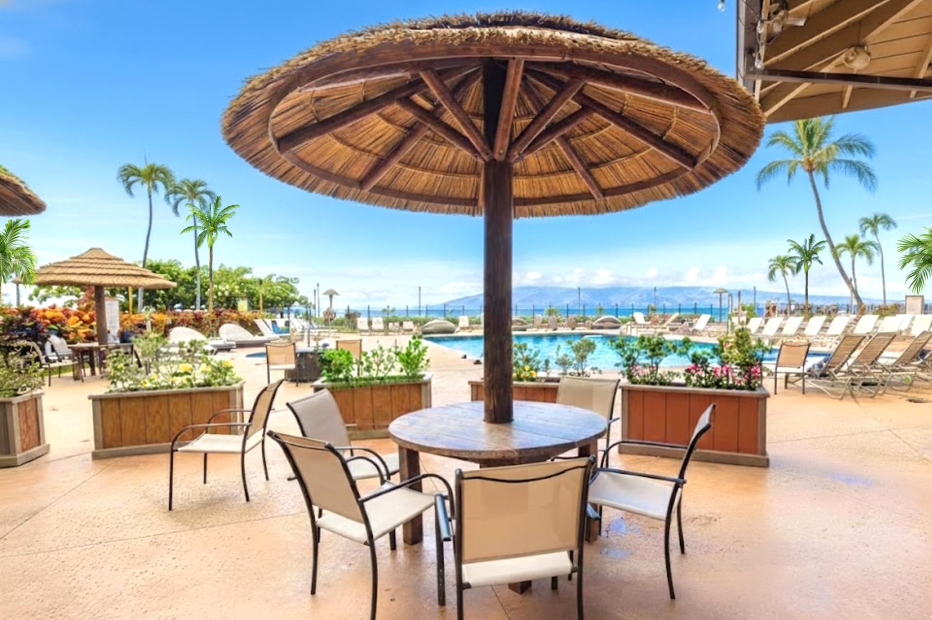 A poolside area with a round table and chairs, a thatched umbrella, and lounge chairs facing a pool.