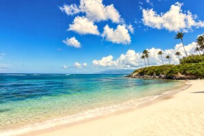 Ein Strand mit klarem türkisblauem Wasser, weißem Sand und Palmen unter blauem Himmel mit Wolken.