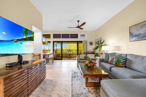 Sala de estar con televisor, sofá, mesa de centro y vistas a una playa a través de amplios ventanales.