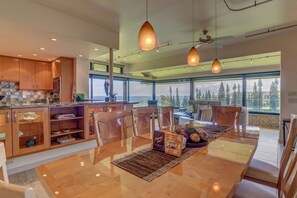 A modern kitchen with wooden cabinets, a dining area with a long table, and large windows offering a view of the outdoors.