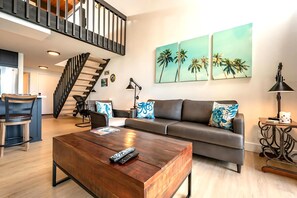 Una sala de estar moderna con una mesa de centro de madera, un sofá gris con cojines decorativos, una escalera de metal negro y una pared con dos cuadros de palmeras.