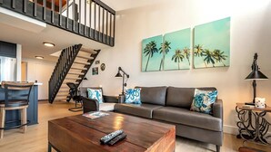 A modern living room with a wooden coffee table, a grey sofa with patterned cushions, a black chair, a staircase, and a wall with three palm tree paintings. - Napili Point | Oceanfront 4 BR, Sleeps 10 | Car Incl. w/ 6+ Nights | NAP ML-1175 by KBM (Lahaina)