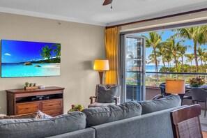 Habitación de hotel con televisor grande, sofá, mesa de comedor con sillas y balcón con vista a palmeras y al océano.