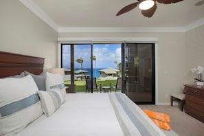 Habitación de hotel con una cama grande, ventilador de techo, puerta corrediza de vidrio que da a un balcón con vista al océano y palmeras.
