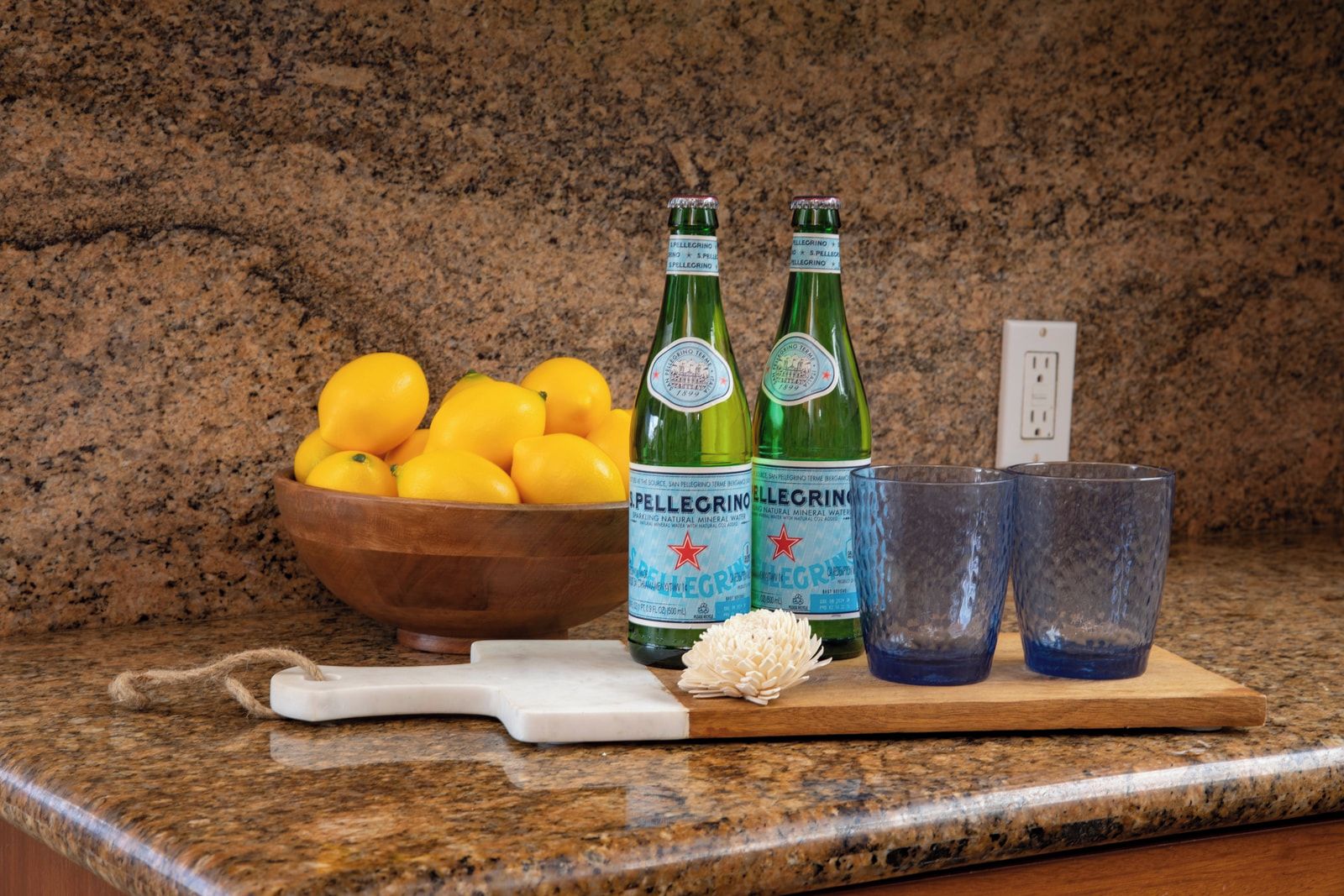 Una encimera de cocina con un bol de limones, dos botellas de agua Pellegrino, dos vasos y una tabla de cortar blanca.