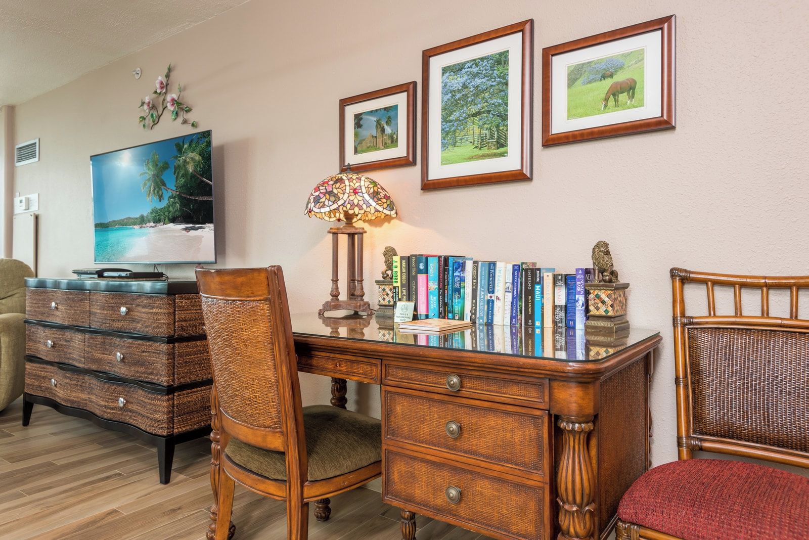A room with a wooden desk, books, a lamp, a television, and framed pictures on the wall.