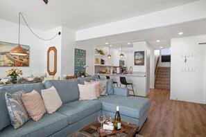 Ein modernes Wohnzimmer mit einer Couch, einem Couchtisch mit Weingläsern, einer Küchenzeile mit Bar und einer Treppe, die eine weitere Ebene hinaufführt.