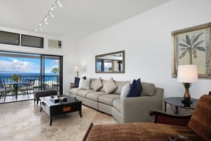 Un salon avec un canapé, une table basse et des fauteuils, un coin repas avec des chaises, un balcon avec vue sur l’océan et un tableau accroché au mur.