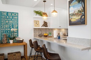 A kitchen with a white countertop, hanging pendant lights, a wooden dining table with chairs, and a decorative sign on the wall. - Kapalua Ridge | Ocean View 3 BR, Sleeps 10 | Car Incl. w/ 6+ Nights | KRV ML-1197 by KBM (Lahaina)