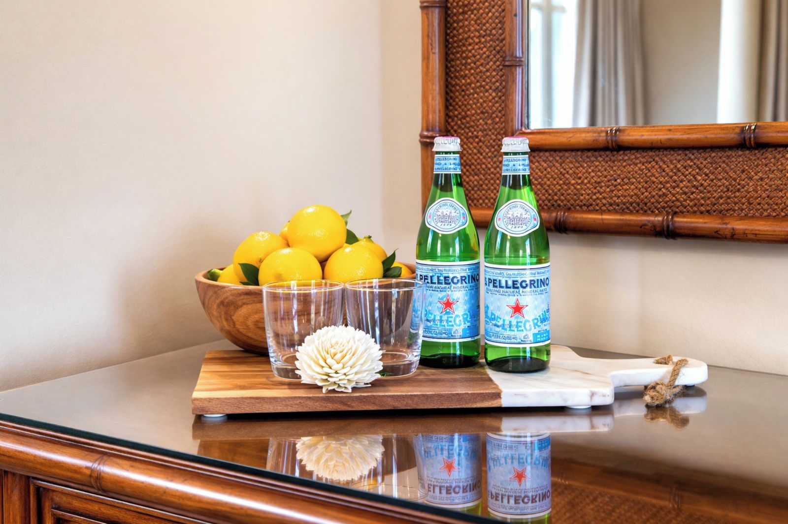 A wooden table with a bowl of lemons, two bottles of Pellegrino water, two glasses, and a decorative flower.