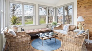A cozy room with wicker furniture, a glass coffee table, and large windows offering a view of a garden. - Farmhouse Retreat, Infrared Sauna, Fire-Pit, Fast Wifi, Golf & 3-Acre Privacy (Valatie)