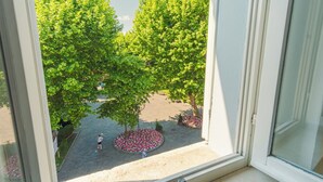 Vista desde una ventana a un patio con árboles, un macizo de flores circular y una zona pavimentada con bancos.