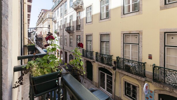 A narrow street with buildings on both sides, featuring balconies with plants and flowers.