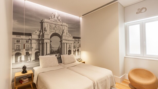 A bedroom with two beds, a large mural of a historical arch, a wooden nightstand, a round chair, and a window with natural light.