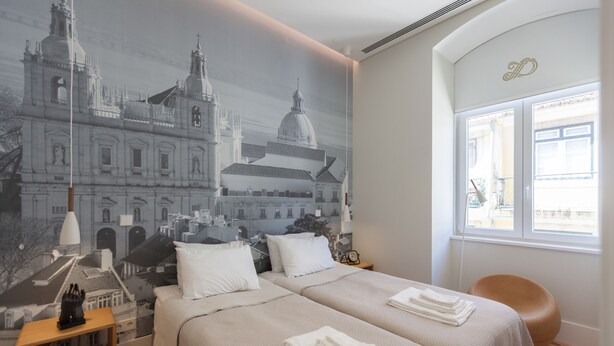 Habitación de hotel con dos camas, un mural de un paisaje urbano, una ventana con vistas y una mesita de noche de madera.