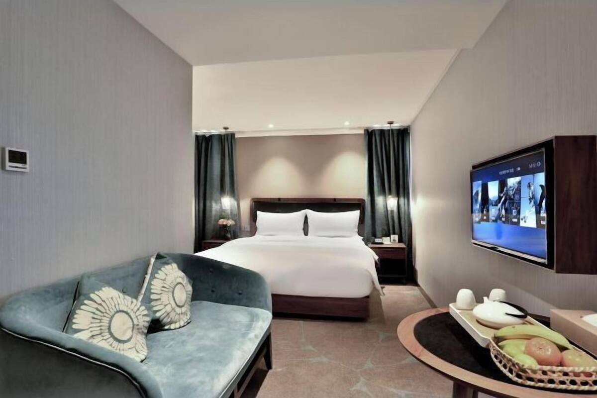 A modern hotel room with a bed, a sofa, a TV, and a small table with fruits.