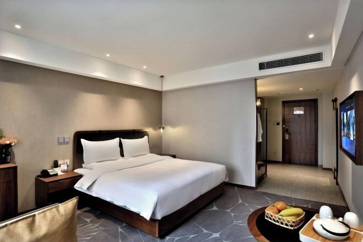 A modern hotel room with a large bed, a nightstand, a desk, and a seating area with fruits and a decorative item.