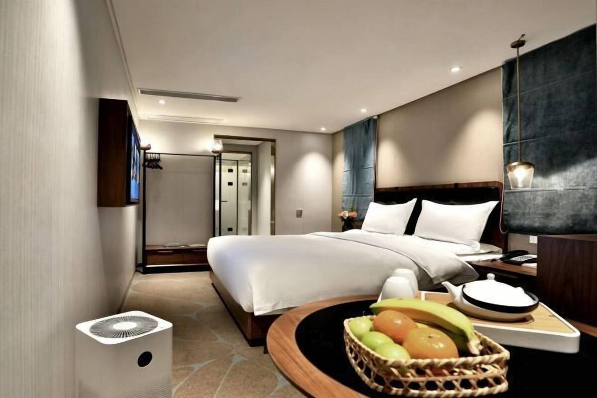 A hotel room with a bed, bedside table, lamp, fruit basket, and a fan.