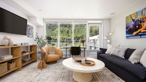 Ein modernes Wohnzimmer mit rundem Couchtisch, einer Couch, einem Fernseher und einem großen Fenster mit Blick auf Bäume.