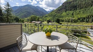Ein Balkon mit rundem Tisch und Stühlen, einem Blumentopf und Blick auf eine gebirgige Landschaft.