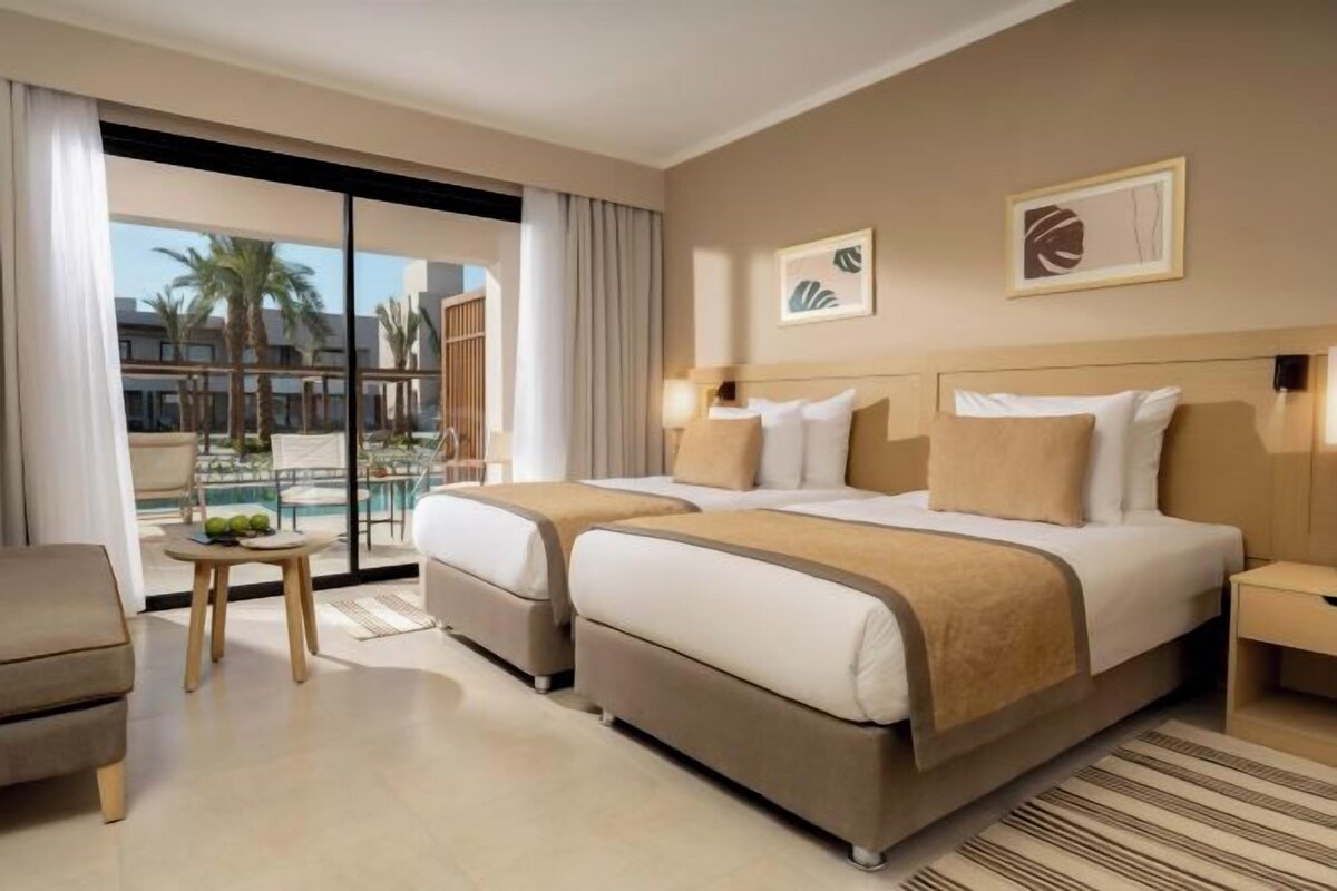 A hotel room with a large bed, a small table with fruits, a chair, and a view of palm trees and buildings.