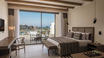 A hotel room with a large bed, a desk, a chair, a small table, a bench, a bedside table, and a view of the city and ocean.