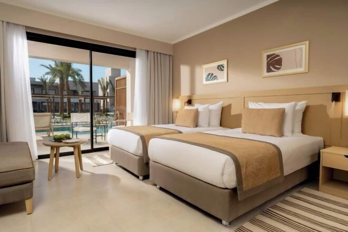 A hotel room with a large bed, a small table with fruits, a chair, and a view of palm trees and buildings.