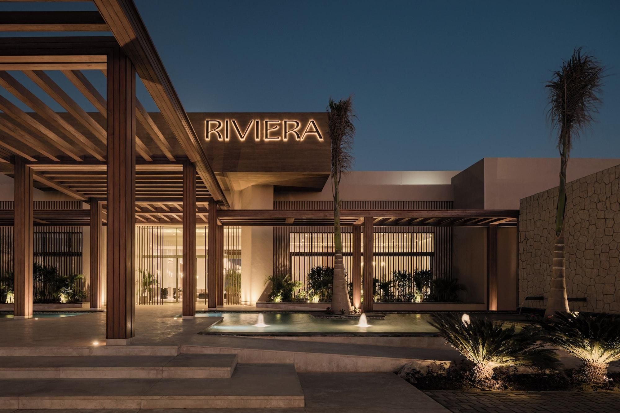 Un edificio moderno con un llamativo letrero 'RIVIERA', una piscina y un área exterior bien iluminada con plantas.