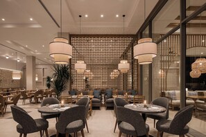 A modern restaurant interior with round tables, grey upholstered chairs, and hanging pendant lights. - JAZ Elite Riviera (Marsa Alam)