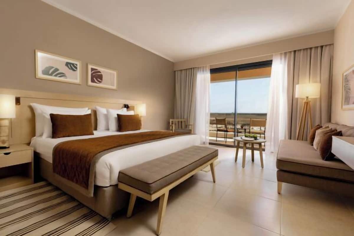 A hotel room with a bed, a bench, a sofa, a dining table, and a view of the outdoors.