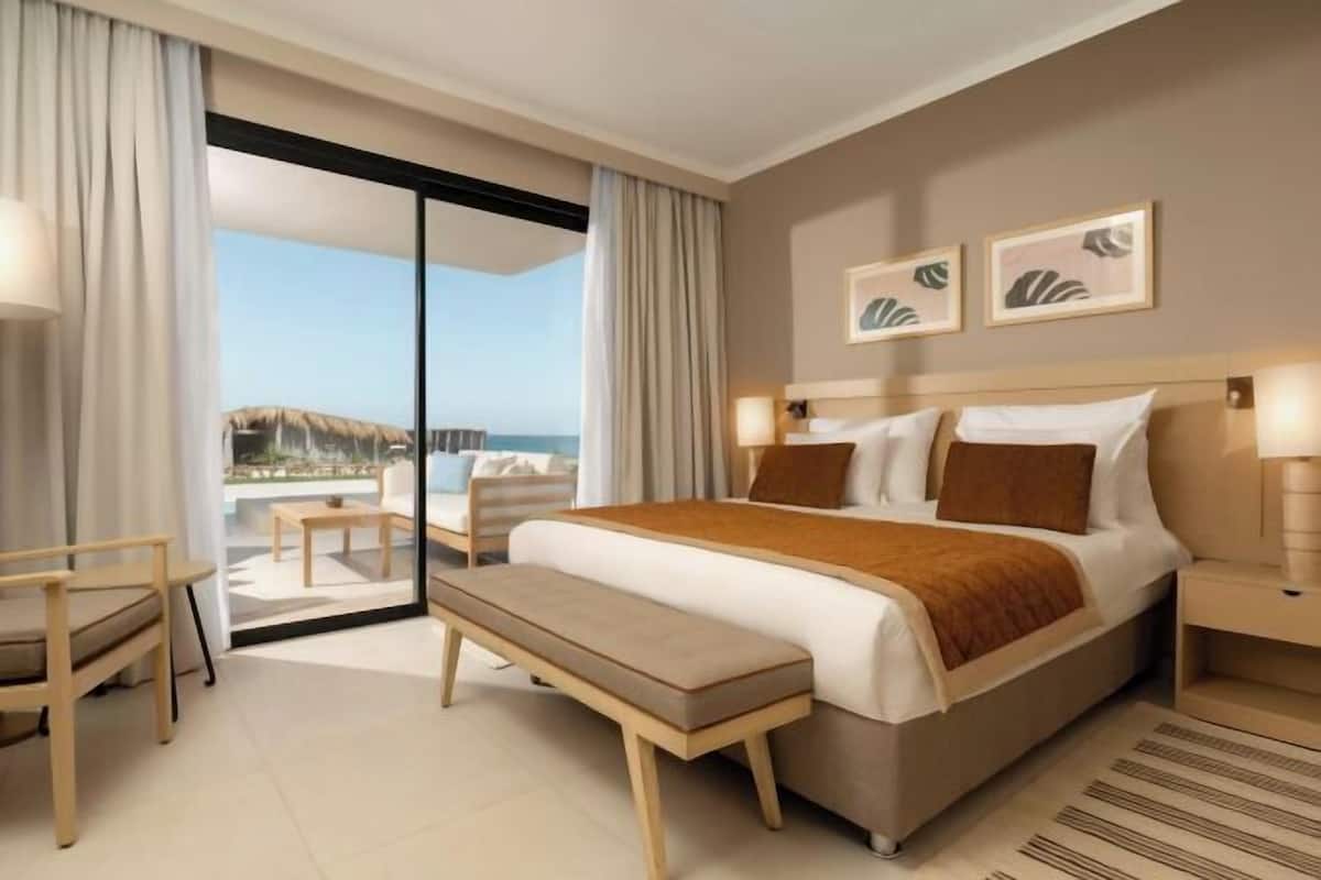 A modern hotel room with a large bed, a bench, a desk, and a view of the ocean.