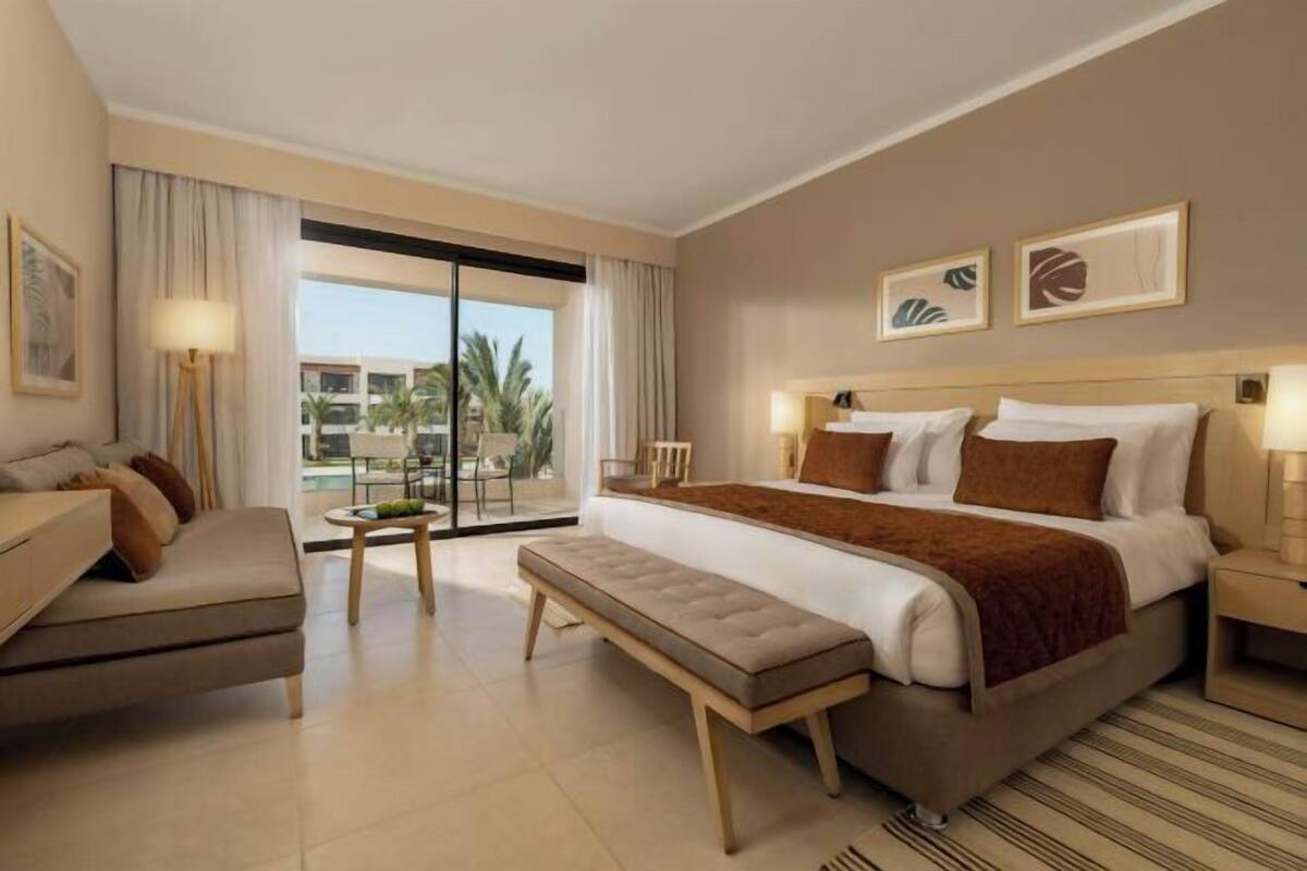 A hotel room with a large bed, a bench, a desk, and a view of palm trees through the window.
