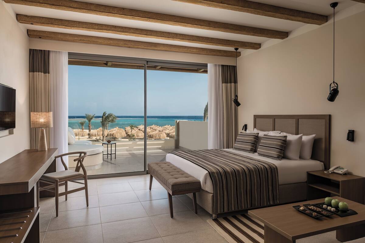 A bedroom with a large bed, a desk, a chair, a bedside table with a phone, and a view of the beach.