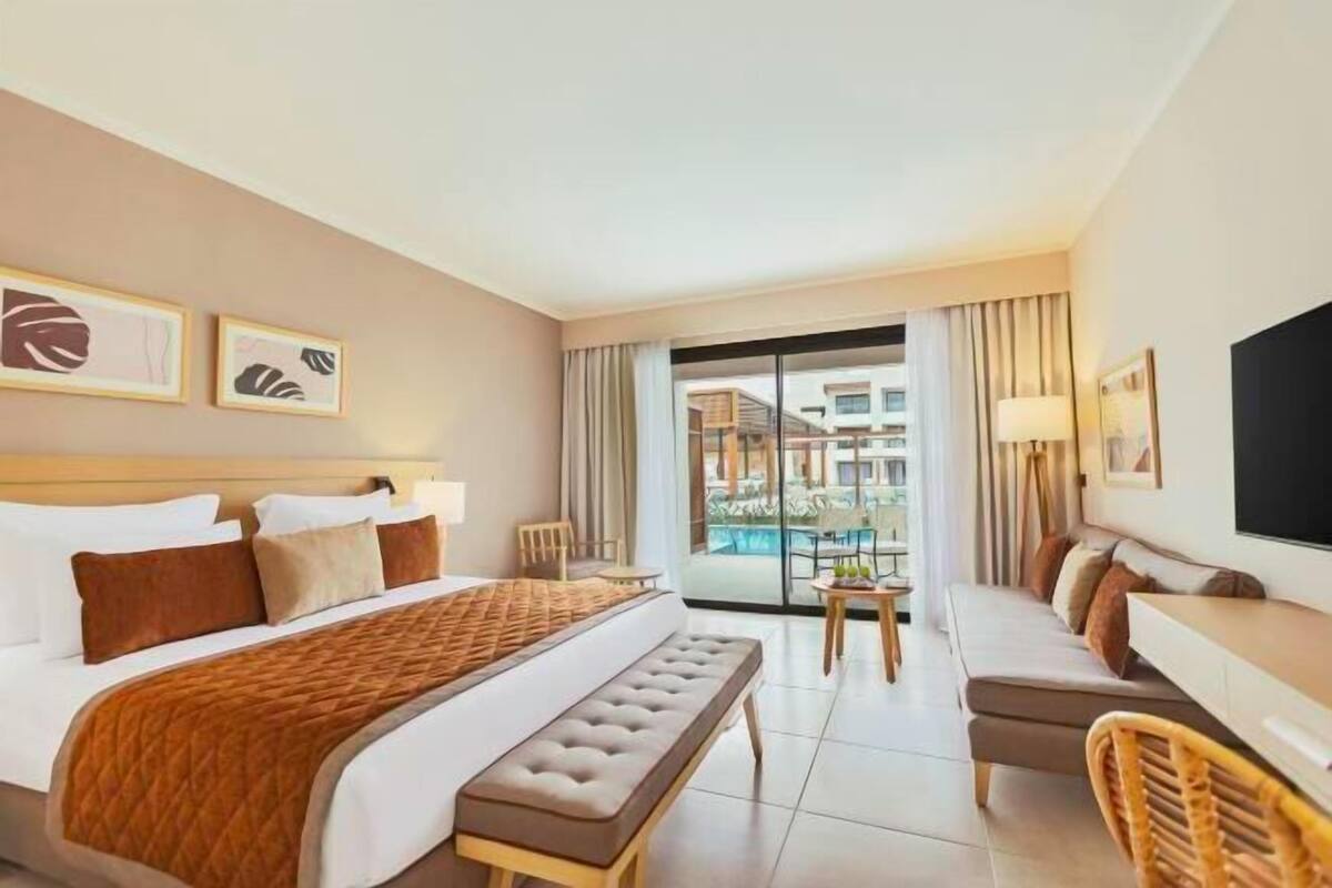 A hotel room with a bed, a bench, a sofa, a TV, and a view of a pool and buildings.
