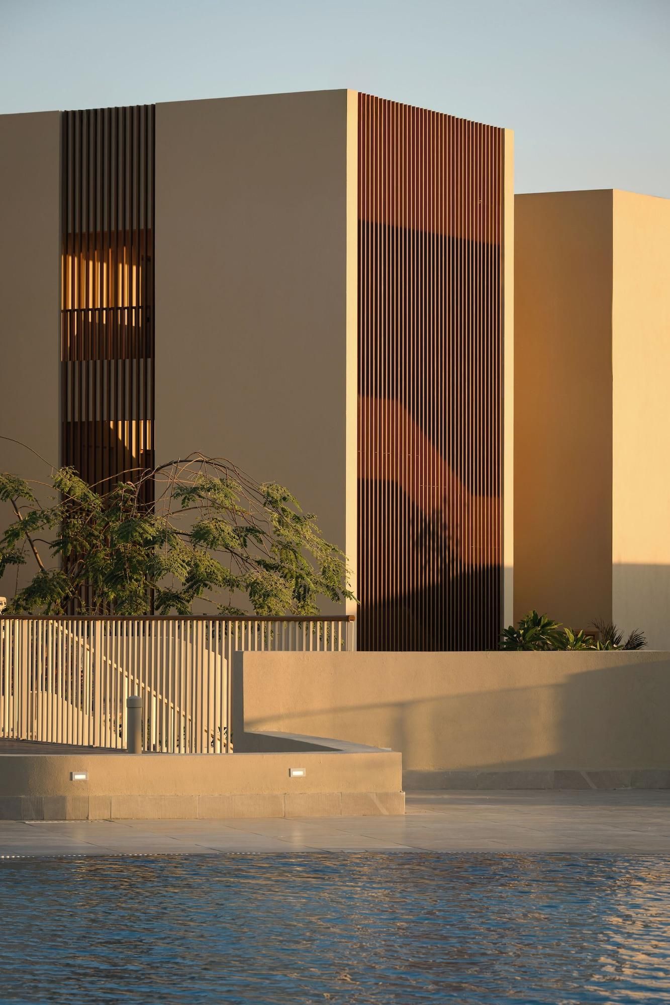 Um edifício moderno com ripas verticais, base de concreto e um espelho d'água na frente.