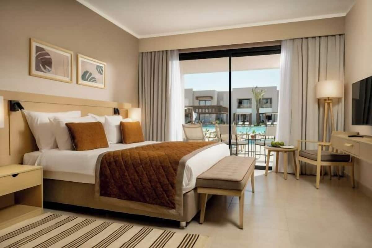 A hotel room with a bed, bedside tables, a desk, a chair, a TV, and a view of a pool and buildings.