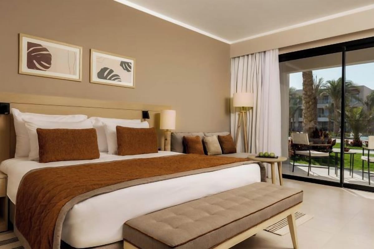 A hotel room with a large bed, a bench, a view of the outdoors, and a balcony with a table and chairs.