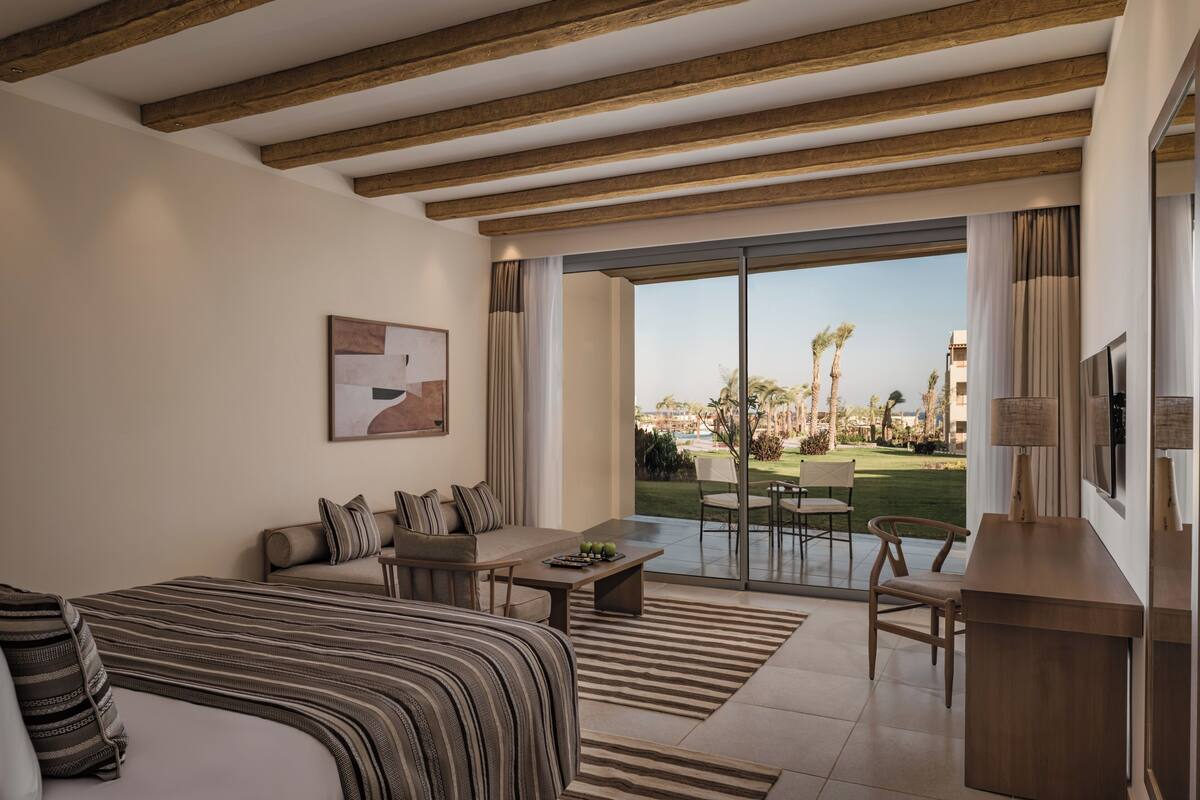 A hotel room with a bed, sofa, dining table, and a view of a garden and buildings.