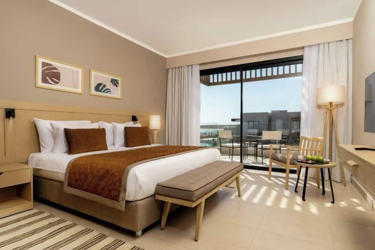 A hotel room with a bed, a bench, a desk, a chair, and a large window.
