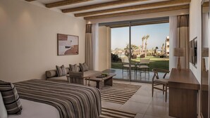 A hotel room with a bed, a sofa, a dining table, and a TV. There is a large window with a view of palm trees and a pool.